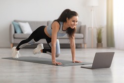 Cheerful active woman doing fitness at home, using laptop, watching online lessons. Positive well-fit lady in sportswear exercising during covid-19 pandemic, watching fitness videos on Internet
