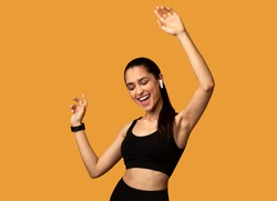 Favourite Song. Portrait Of Excited Young Sportive Lady Wearing Wireless Earbuds Listening To Music And Dancing Isolated Over Orange Studio Background. Lady With Closed Eyes Enjoying Playlist