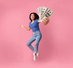 Full length portrait of a joyful young asian woman jumping up high and holding bunch of money banknotes, showing close to camera, celebrating win, isolated over pink studio background, collage