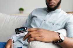 Unrecognizable Black Man With Pulse Oximeter On Hand Measuring Oxygen Saturation Level At Home. Pulseoxymeter Medical Device, Pulseoxymetry Clip Machine Monitorin Ox Rate. Cropped, Selective Focus