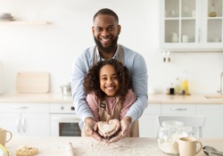 Bonding And Togetherness Concept. Portrait of happy african american dad and daughter baking in the kitchen and holding dough in heart shape in hands, preparing present for mother's day