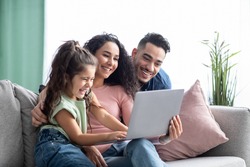 Cheerful arabic family having fun with laptop at home, watching photos together. Middle Eastern parents and their little daughter using computer while sitting on couch in living room, copy space