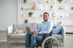 Happy disabled black man in wheelchair using laptop, working online from home, copy space. Millennial African American handicapped guy having business or studying on web