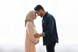 Loving pregnant muslim couple bonding together at home, standing near window, expecting arab family touching foreheads and smiling, romantic husband embracing belly of his wife in hijab, side view