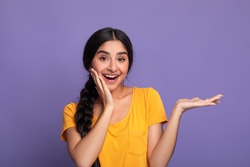 Wow, Look Here. Portarit of smiling young indian lady holding outstretched hand with open palm, advertising and showing free empty space and touching her cheek posing isolated on purple studio wall