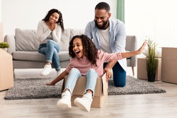 New Home, People And Real Estate Concept. Excited African American family having fun and celebrating moving day, cheerful father riding his little daughter in cardboard box container in living room