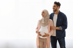Offer For Future Parents. Portrait Of Pregnant Religious Muslim Couple Standing Near Window At Home, Looking Aside At Copy Space With Happy Excitement, Smiling Arab Expectant Woman Wearing Hijab