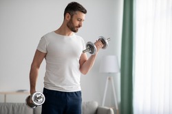 Closeup of handsome middle-aged man doing dumbbell workout at home, working on arms strength, looking at his biceps, copy space. Athletic man lifting dumbells up over living room interior