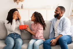 Portrait of African American girl making surprise for her mum, giving present, greeting woman with Mother's day or birthday. Smiling lady covering eyes sitting on the couch in living room