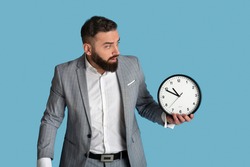 Scared office employee with clock missing deadline on blue studio background. Millennial bearded businessman having time management problem, being late for important meeting