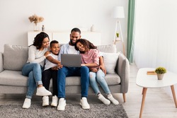 Family Weekend. Portrair of cheerful African American parents and their little children using laptop at home together, watching movie or browsing internet, sitting on the couch in living room
