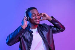Music Lover. Handsome Black Guy In Wireless Headphones And Leather Jacket Listening His Favorite Songs, Relaxed African American Man Standing With Closed Eyes Under Neon Light Over Purple Background