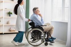 Side view of young doctor helping elderly disabled man in wheelchair, indoors. Nurse taking care of handicapped male patient at home, providing medical assistance to impaired senior adult