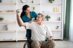 Rehabilitation of disabled people concept. Young physiotherapist helping senior male patient in wheelchair exercise at home. Handicapped elderly man training with dumbbells