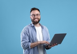 Cheerful millennial guy in glasses using laptop computer for online work or communication on blue studio background