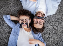 Funtime together. Curly father and son playing with fake moustache on sticks, lying on floor, top view