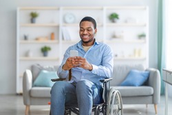 Disabled black man in wheelchair using smartphone, browsing web or watching movie at home. Handicapped young guy checking social media, speaking to friend on mobile device