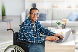 Cheerful handicapped black man in wheelchair using laptop for online job from home, having business meeting on web. African American disabled freelancer working on remote project
