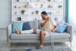 Young African American guy with plastered leg sitting on sofa, leaning on crutches at home. Millennial black man suffering from pain in broken limb, resting on couch, full length portrait