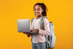 Portrait of smiling African American girl holding and using laptop computer, wearing backpack, typing on keyboard, posing and looking at camera standing isolated over yellow studio background