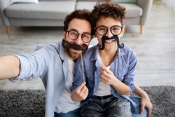 Funny selfie. Happy father and son taking selfie, holding fake moustache on sticks, wearing glasses and smiling at camera, having fun at home, sitting on floor carpet. Parenthood concept