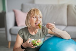 Sports and nutrition concept. Smiling senior lady leaning on fitness ball, eating fresh vegetable salad at home. Athletic mature woman enjoying her diet and exercising program