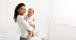 Young Black Mom Holding Baby Toddler, Hugging Carrying Adorable Little Son Posing Standing Indoors. Child Care, Motherhood And Maternity Leave Concept. Panorama, Empty Space For Text