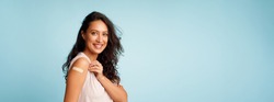 Coronavirus Vaccination Advertisement. Happy Vaccinated Woman Showing Arm With Plaster Bandage After Covid-19 Vaccine Injection Posing Over Blue Background, Smiling To Camera. Panorama, Blank Space