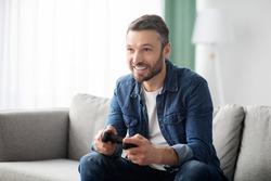 Joyful middle-aged bearded man playing video game with joystick at home, sitting on couch in living room, having fun at weekend, copy space. Home entertainment, leisure, domestic lifestyle