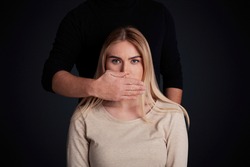 Tyranny, psychological pressure and discrimination concept. Male in dark clothes closes mouth of frightened young attractive female, isolated on black background, free space, studio shot