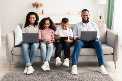 Modern Weekend With Family. Portrait of smiling African American parents and their little children holding and using different electronic gadgets while sitting on the sofa in living room at home
