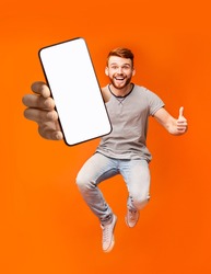 Happy redhead man jumping up in air, showing thumbs up gesture, demonstrating smartphone with empty screen, recommending new mobile application on orange studio background