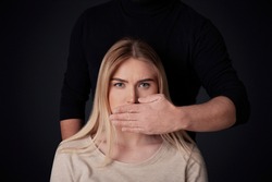 Domestic violence, tiran, sexism, abuse and shut up. Male abuser in dark clothes closes mouth to sad young woman making impossible to talk, isolated on black background, free space, studio shot
