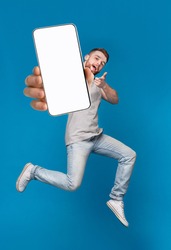 Emotional millennial redhaired guy pointing finger at camera, jumping with mobile phone on blue studio background, mockup for mobile app on white screen. Creative collage with website template