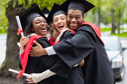 Black guy and two young ladies in graduation costumes posing at camera at university campus, holding diplomas, laughing and hugging, having happy graduation day, closeup portrait