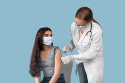Medical doctor or nurse giving coronavirus vaccine shot to Indian teen girl over blue studio background. Female teenager in face mask getting covid-19 vaccine, participating in immunization campaign
