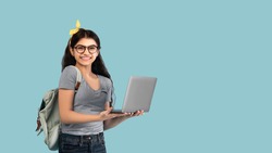 Portrait of smiling Indian teen girl using laptop computer, studying online on blue studio background, banner design with copy space. Happy teenager lady participating in educational webinar