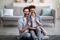 Father best example of success and courage. Son and dad holding fake moustache on sticks and smiling to camera, having fun at home, sitting on floor in living room