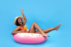 Positive black woman sitting on inflatable ring, holding tropical cocktail on blue background, full length. Happy African American lady having beach party with pool float and refreshing summer drink