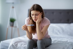 Concerned young woman upset with pregnancy test results, sitting on bed at home with worried face. Millennial lady not ready to become mother or being unable to conceive baby