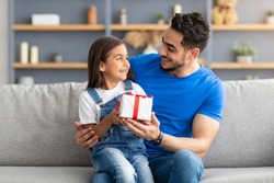 This Present Is For You. Portrait of loving girl sitting on dad's lap and greeting him with father's day or birthday, holding wrapped gift box, happy family celebrating holiday together at home