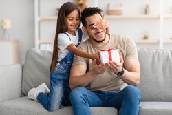 Portrait of cute little girl holding and giving wrapped gift box, making surprise for her excited dad, covering his eyes, greeting young man with father's day or birthday, celebrating at home