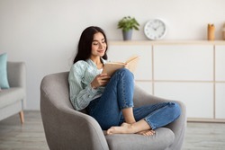 Stay home pastimes. Millennial Indian woman sitting in cozy armchair with open book indoors. Beautiful young lady reading exciting story, enjoying lazy morning, having relaxing weekend