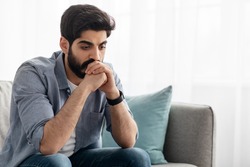 Male depression. Upset arab man having problems, sitting on couch at home and thinking, having heavy thoughts, breake up with lover or financial difficulties concept, empty space