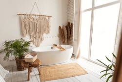 Cozy modern minimalistic bathroom interior with big white bathtube and window, green plants and rustic decoration elements indoors. Light empty bathing room background, design ideas. No people