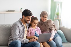 Young muslim parents and girl using laptop together at home, surfing internet or having video chat with someone, sittig on sofa. Happy arab man, child and woman in hijab staying home on isolation
