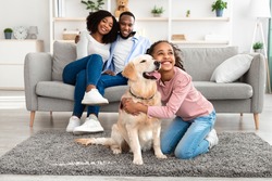 Man's Best Friend. Portrait of smiling black girl hugging her happy dog in living room at home, looking away, cheerful parents sitting on the couch and looking at daughter playing with pet