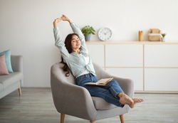 Gorgeous Indian woman stretching in comfy armchair while reading indoors. Attractive young lady with open book, enjoying lazy morning, having peaceful weekend. Stay home hobbies