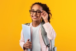 Education Concept. Portrait of positive smiling African American girl touching glasses holding textbooks, wearing backpack and looking at camera isolated on yellow studio background
