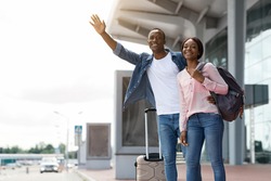 Portrait Of Happy African American Couple Catching Taxi Outdoors Near Airport Terminal After Arrival, Smiling Young Black Travellers Carrying Luggage, Man Raising Hand To Stop Cab, Copy Space